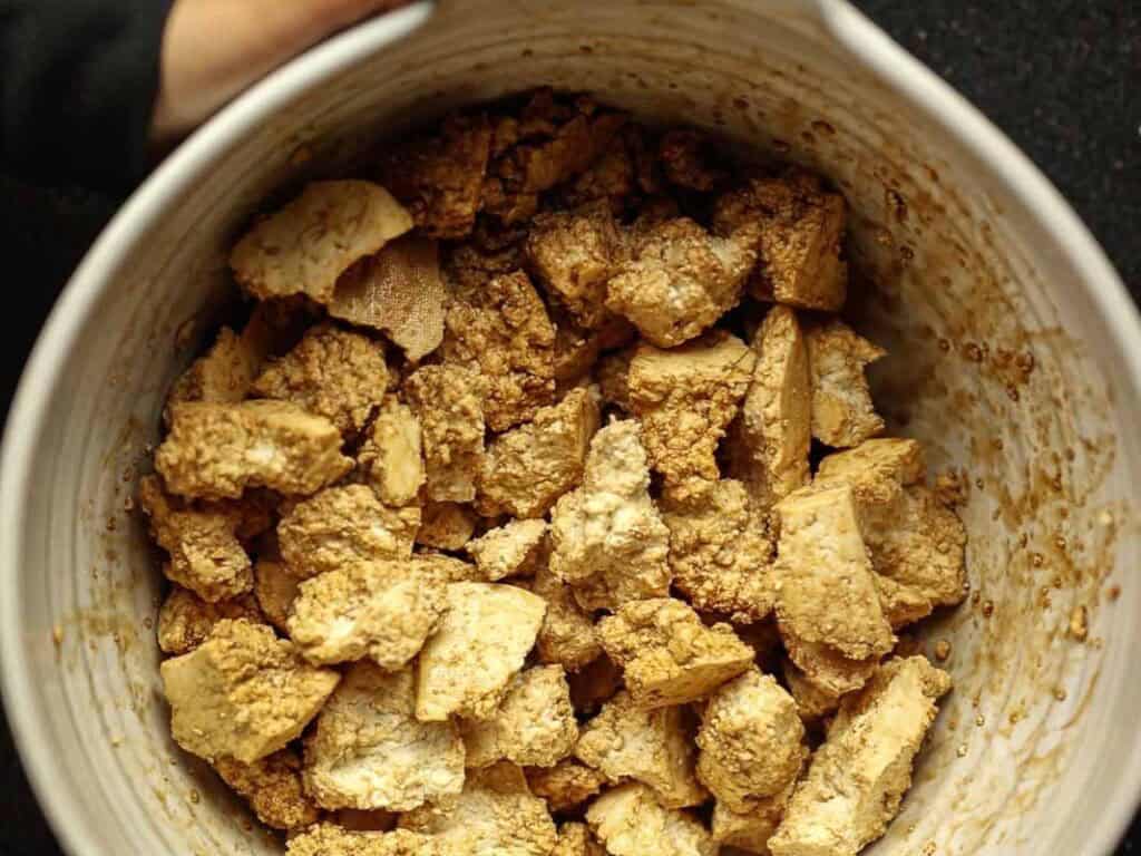 A large bowl filled with chunks of super firm tofu marinating in a brown sauce. The tofu pieces are irregular in shape, and the marinade adds a textured, glazed appearance. One hand is partially visible holding the bowl.